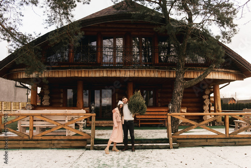 Young beautiful happy lovers man and woman, love story in winter with a live Christmas tree in their hands on the background of a wooden house © ksyusha_yanovich