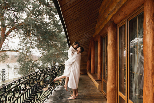 Young beautiful happy newlyweds in love a man and a woman in white bath robes on the balcony of the house smile and hug