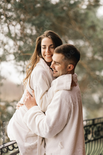 Young beautiful happy newlyweds in love a man and a woman in white bath robes on the balcony of the house smile and hug