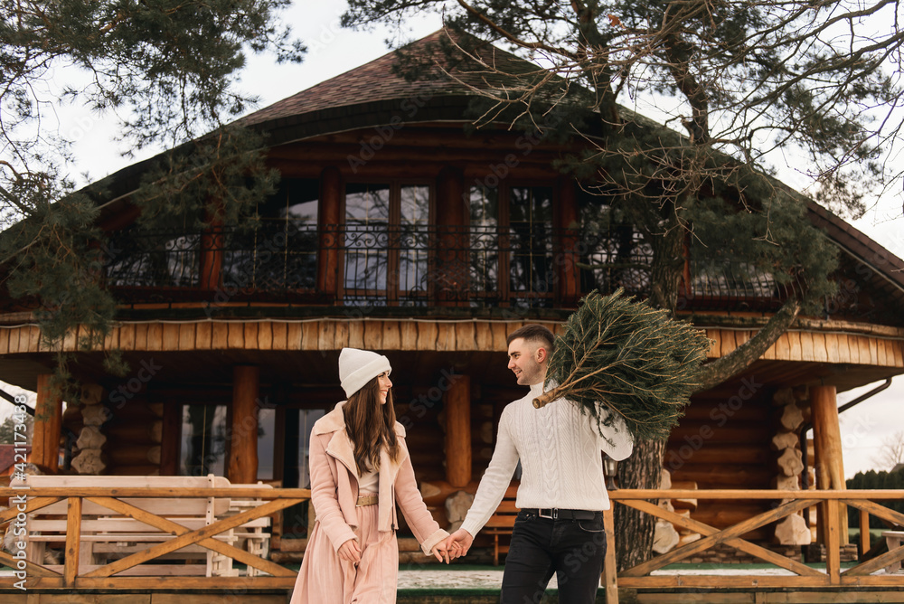 Young beautiful happy lovers man and woman, love story in winter with a live Christmas tree in their hands on the background of a wooden house