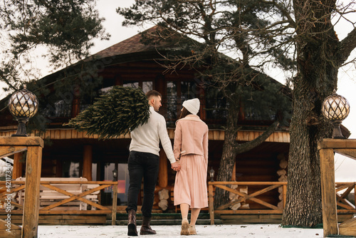 Young beautiful happy lovers man and woman, love story in winter with a live Christmas tree in their hands on the background of a wooden house © ksyusha_yanovich