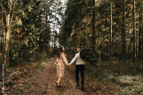 Young beautiful happy lovers man and woman, love story in winter with a live Christmas tree in a pine forest in the sun