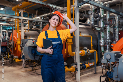 The concept of industrial production. A young smiling worker in a work uniform and taking off her helmet, with a tablet in her hand. In the background is a boiler room