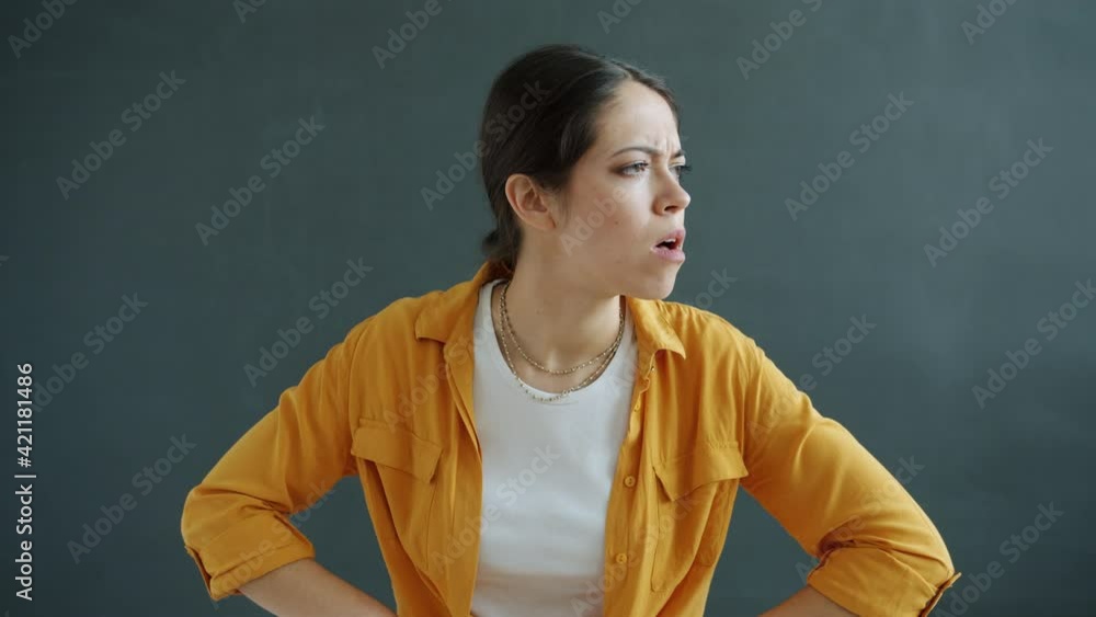 Portrait of unhappy and angry young woman looking at camera standing akimbo on gray background. Nonverbal communication and bad mood concept. Portrait of unhappy and angry young woman looking at camera standing akimbo on gray background. Nonverbal communication and bad mood concept.