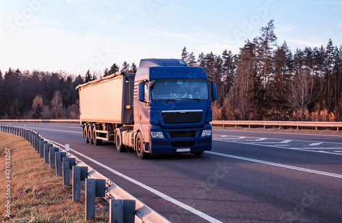 A modern truck transports bulk cargo along an asphalt road against the backdrop of a forest and sky. Concept for the transportation of solid bulk cargo in the form of grain in special trailers photo