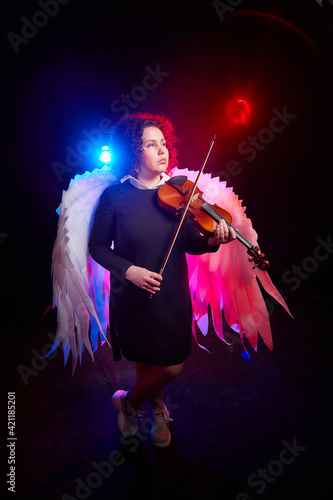 A brunette curly girl in black dress looking as hipster or teenager with violin and white angel wings on a black background. Model, violinist, actress posing in the studio