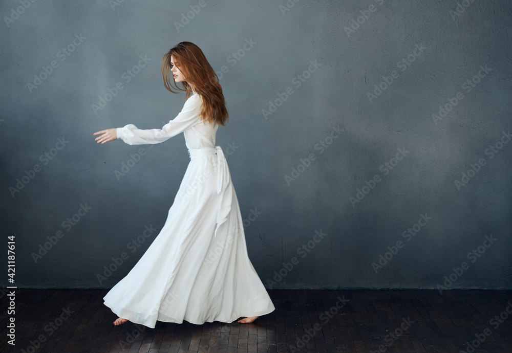 Romantic woman with red hair dances on a gray background in a white dress Romantic woman with red hair dances on a gray background in a white dress