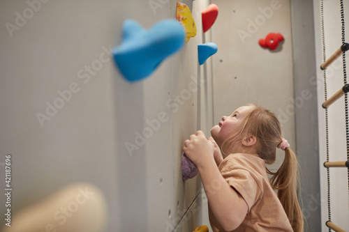 Fototapeta Naklejka Na Ścianę i Meble -  Side view portrait of cute little girl with down syndrome climbing wall and looking up while exercising in sports set at home, copy space