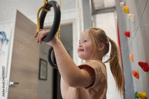 Fototapeta Naklejka Na Ścianę i Meble -  Side view portrait of cute girl with down syndrome enjoying exercise at home in sports set with rings, copy space