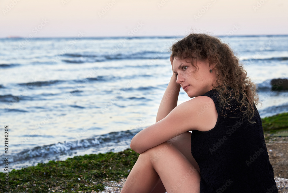 A thoughtful woman sits alone on the sea beach near the water.