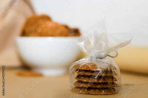 In the foreground are five sesame cookies wrapped in cellophane. In the background there is a white plate and a brown paper bag out of focus. Homemade baking.
