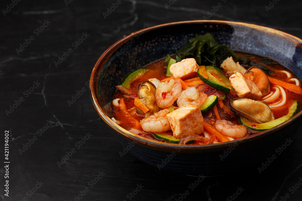 Asian ramen soup with shrimps, mussels, tofu, carrot, zucchini and seaweed. Dish isolated in a dark blue bowl, close-up on a black marble background. Asian cuisine. Copy space. Asian ramen soup with shrimps, mussels, tofu, carrot, zucchini and seaweed. Dish isolated in a dark blue bowl, close-up on a black marble background. Asian cuisine. Copy space.