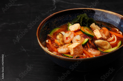Asian ramen soup with shrimps, mussels, tofu, carrot, zucchini and seaweed. Dish isolated in a dark blue bowl, close-up on a black marble background. Asian cuisine. Copy space.