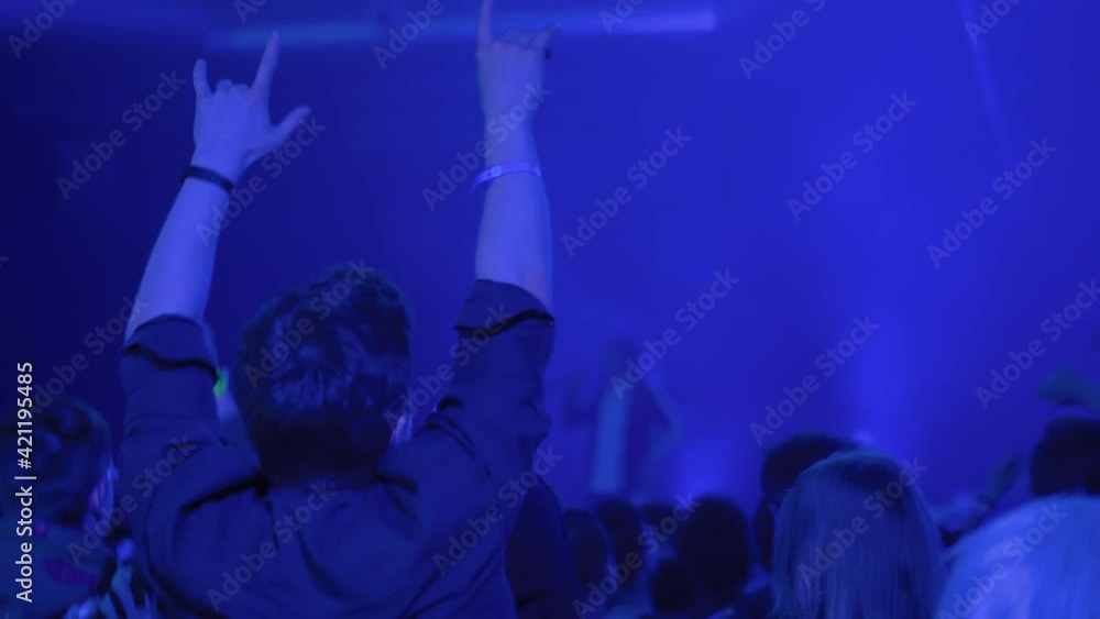 Slow motion: teenage boy partying, cheering, raising hands up and jumping at rock concert in front of stage of nightclub. Bright colorful blue stage lighting. Nightlife and entertainment concept Slow motion: teenage boy partying, cheering, raising hands up and jumping at rock concert in front of stage of nightclub. Bright colorful blue stage lighting. Nightlife and entertainment concept