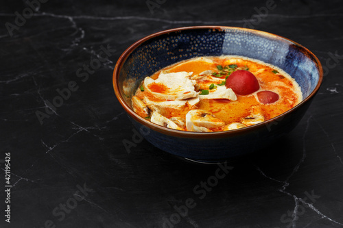 Asian chicken curry soup with chicken breast, tomatoes, mushrooms and spring onion. Close-up photo of the dish served in a dark blue bowl, isolated on a black marble background. Asian cuisine.