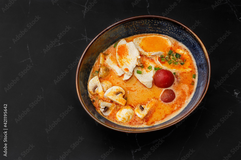Asian chicken curry soup with chicken breast, tomatoes, mushrooms and spring onion. Close-up photo of the dish served in a dark blue bowl, isolated on a black marble background. Asian cuisine.