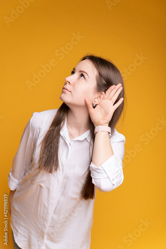 Young woman on isolated yellow background shows that she is hard of hearing by putting her palm to her ear