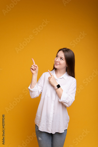 Young woman on an isolated yellow background shows at an imaginary copyspace to insert an advertisement, looks in upward direction