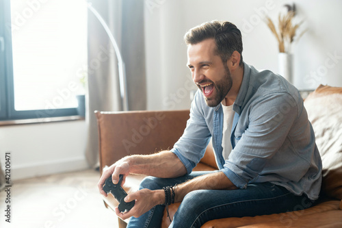Portrait of young bearded hipster guy sitting on the couch in the living room, holding joystick, screaming, and winning a video game match, male addicted gamer with an open mouth is about to succeed