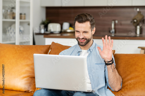 Front view of smiling young bearded hipster guy sitting on the couch, waving at laptop with high five, video-calling with family coworkers online, studying remotely, male freelnacer talking on webcam photo