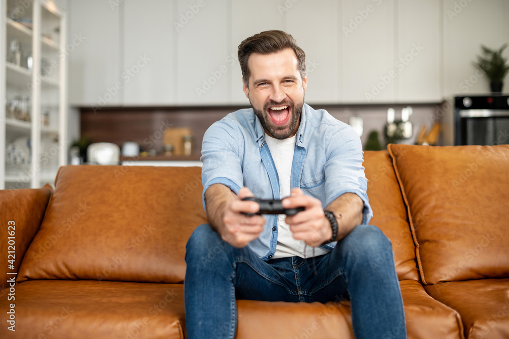 Front view of young bearded hipster man sitting on the couch in the living room staying at home and playing, holding wireless joystick, screaming, and winning intense video game match, victory concept Front view of young bearded hipster man sitting on the couch in the living room staying at home and playing, holding wireless joystick, screaming, and winning intense video game match, victory concept
