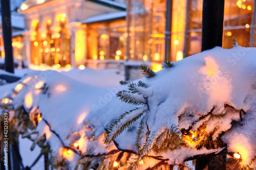 Obraz na płótnie Snow-covered Christmas decorations with spruce branches and Christmas garlands on the street. Beautiful classic European architecture in the background. w salonie