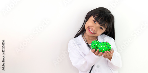 Asian child girl in a white doctor's uniform holds a green ball that looks like a virus or bacteria in her hand and a smiling face.