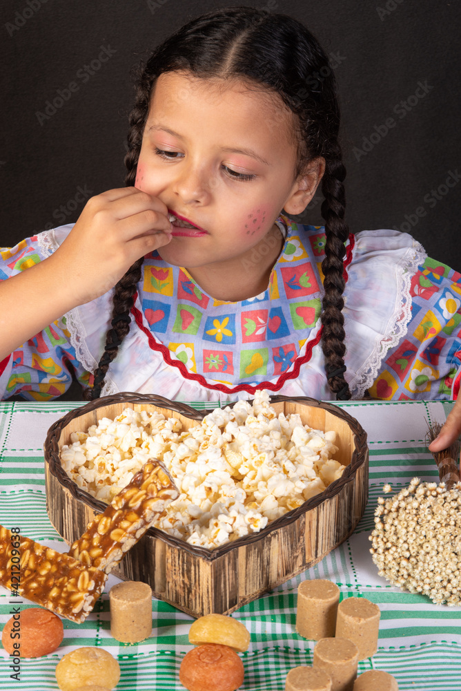 Festa junina in Brazil, brazilian girl with braids and dressed for festa junina with popcorn and typical sweets, black background, selective focus. Festa junina in Brazil, brazilian girl with braids and dressed for festa junina with popcorn and typical sweets, black background, selective focus.