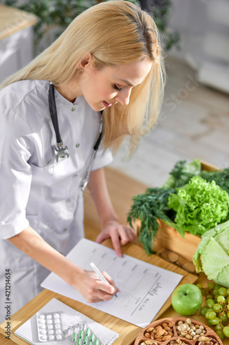 Top view on female Doctor standing by table with fresh raw fruit and vegetables and writing individual weight loss plan