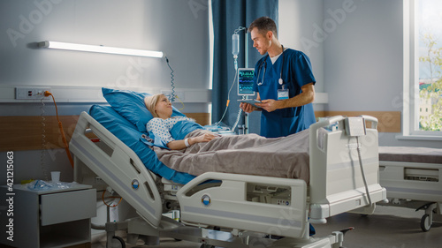 Hospital Ward: Friendly Male Nurse Talks to Beautiful Female Patient Resting in Bed. Male Nurse or Physician Uses Tablet Computer, Does Checkup, Woman Recovering after Successful Surgery 