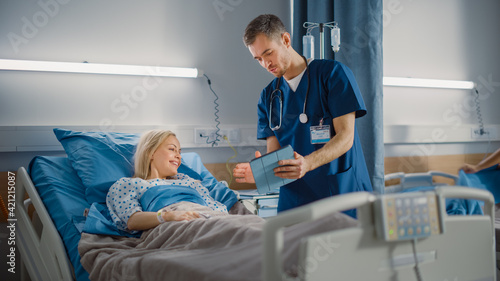 Fotografering Hospital Ward: Friendly Male Nurse Talks to Beautiful Female Patient Resting in Bed