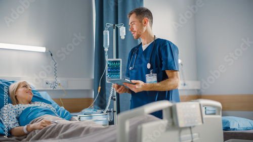 Hospital Ward  Friendly Male Nurse Talks to Beautiful Female Patient Resting in Bed. Male Nurse or Physician Uses Tablet Computer  Does Checkup  Woman Recovering after Successful Surgery 