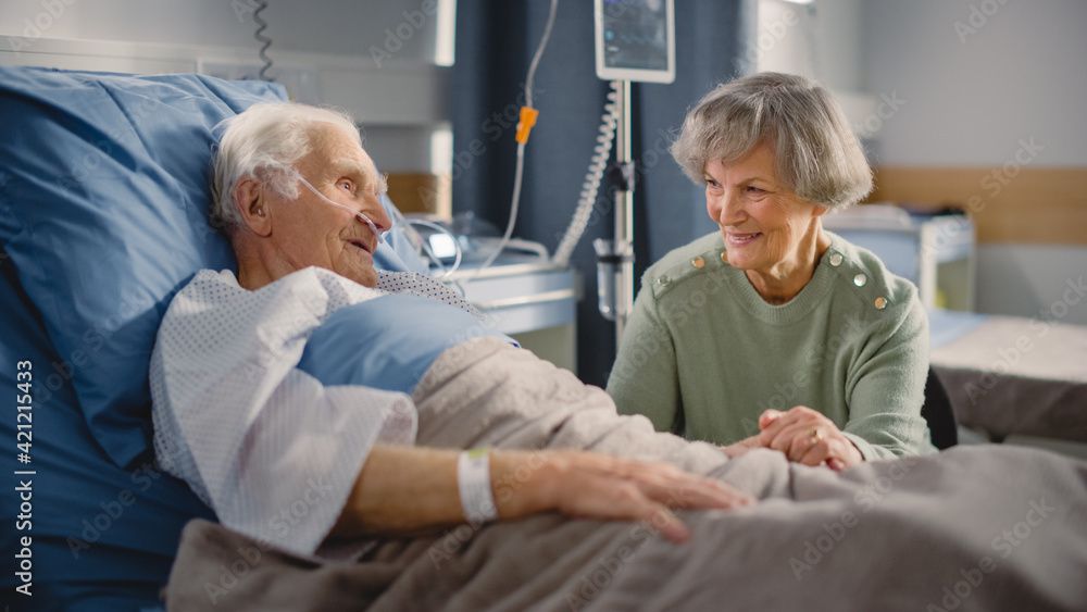 Fototapeta premium Hospital Ward: Elderly Man Resting in Bed, His Caring Beautiful Wife Supports Him Sitting Beside, Happy together They Talk about Good Times. Old Man Fully Recovering after Successful Surgery