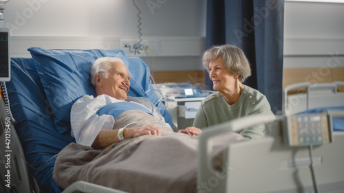 Hospital Ward  Elderly Man Resting in Bed  His Caring Beautiful Wife Supports Him Sitting Beside  Happy Couple Together  They Talk  Remember Good Times. Old Man Recovering successfuly After Sickness