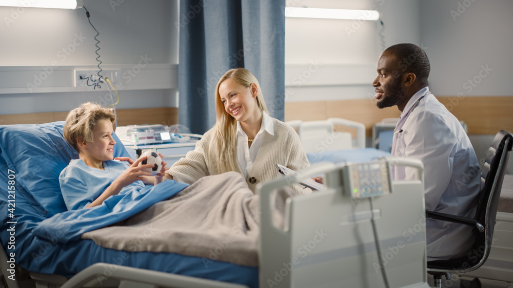 Fototapeta premium Hospital Ward: Handsome Young Boy Resting in Bed with Caring Mother Visits to Support Him, Friendly Doctor Talks. Smiling Patient Fully Recovering after Sickness or Successful Surgery