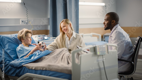 Hospital Ward: Handsome Young Boy Resting in Bed with Caring Mother Visits to Support Him, Friendly Doctor Talks. Smiling Patient Fully Recovering after Sickness or Successful Surgery