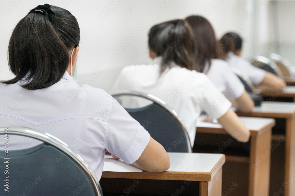 Rear view of the teenage college students sit on lecture chairs do final examination and write on examination paper answer sheets in the classroom. University students in uniform in classroom. Rear view of the teenage college students sit on lecture chairs do final examination and write on examination paper answer sheets in the classroom. University students in uniform in classroom.