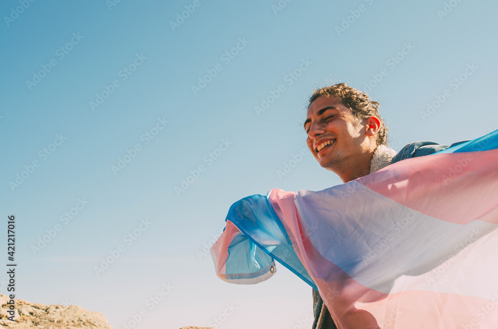 Happy Young Man Smiling While Holding Transgender LGTB Flag With Open Arms and Blue Sky Background - obrazy, fototapety, plakaty 