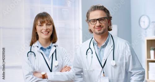 Portrait of Caucasian joyful team of doctors male and female colleagues in white coats standing in cabinet in clinic and smiling to camera. Professional physicians looking at camera  hospital concept