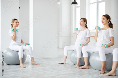 Group of pregnant females sit on fitballs training with dumbbells