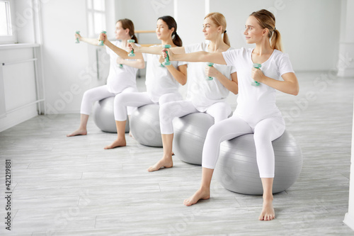 group of women in sportswear doing pregnant exercises sitting on fitness sword
