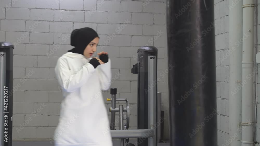 confident Muslim woman stands in the pose of a fighter, boxing in the gym, boxing in the fitness center, a woman dressed in a white hijab. Muslim woman hits a punching bag. confident Muslim woman stands in the pose of a fighter, boxing in the gym, boxing in the fitness center, a woman dressed in a white hijab. Muslim woman hits a punching bag.