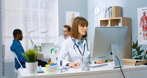 Caucasian young female physician working in medical center sitting at desk typing on computer at workplace while male colleague speaking with African American patient on background  medic concept