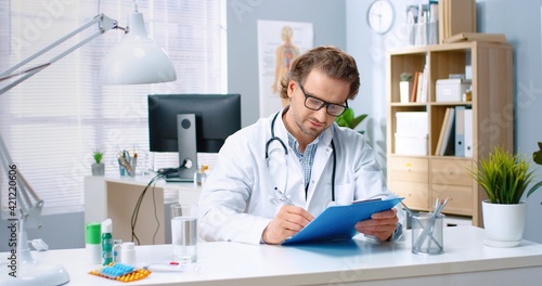 Portrait of middle-aged happy positive handsome Caucasian male doctor in glasses working in hospital cabinet writing while sitting at desk  looking at camera and smiling in good mood  medic concept