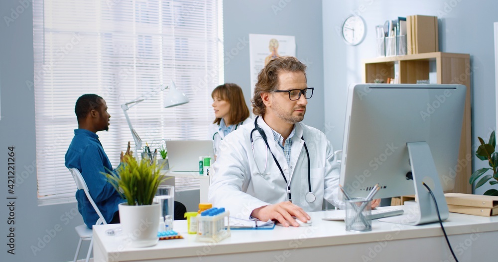 Portrait of happy bearded Caucasian middle-aged male doctor working in hospital cabinet typing on computer while female physician talking with African American patient on background
