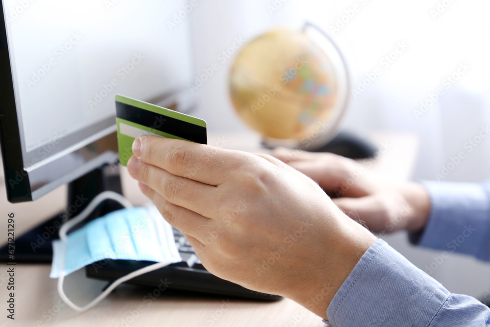 Fototapeta premium Man holding credit bank card types on PC keyboard. Concept of online shopping and payment, buying a travel tour during coronavirus pandemic