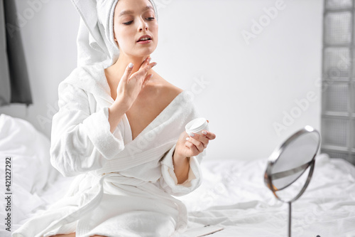 attractive female in towel and bathrobe applying cream on body in the morning