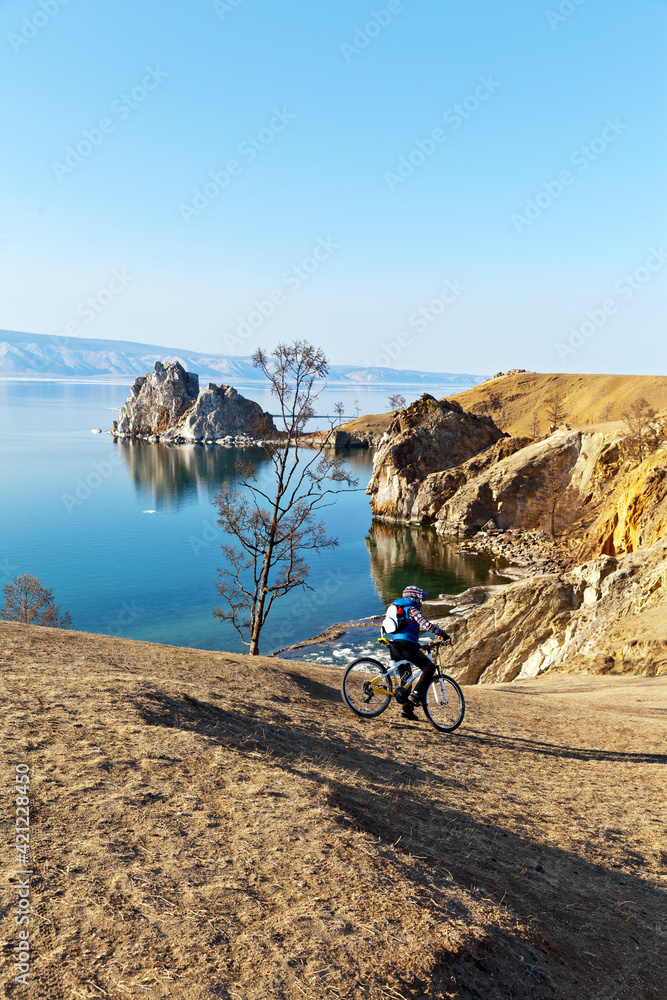 Fototapeta premium Beautiful spring landscape of Baikal Lake on sunny May day with a view on the famous Shamanka Rock and the rocky coast of Olkhon Island. Girl tourist rides a bike on the coastal hills. Active vacation