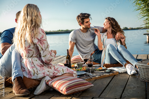 Friends having fun on picnic near a lake, sitting on pier eating and drinking wine.	