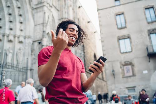 Cheerful travel blogger with curly hair using smartphone gadget for social networking during journey vacations for visiting historic city, smiling hipster guy connecting to 4g internet for browsing